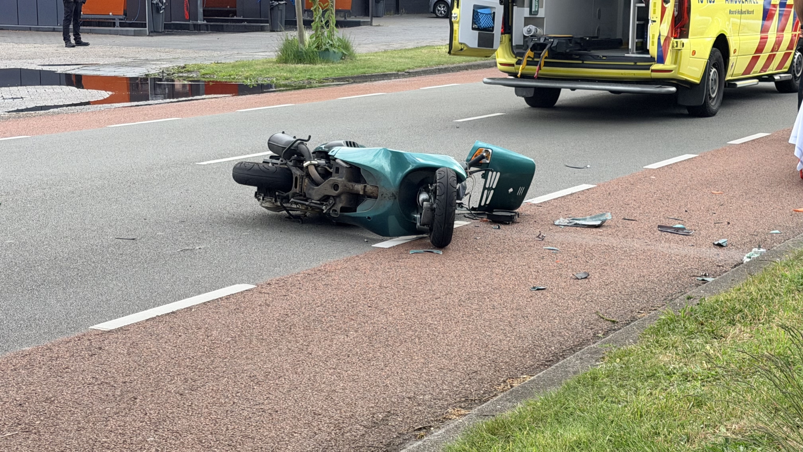 Hulpdiensten helpen een gewonde persoon op een brancard bij een ongeval op straat; een ambulance staat klaar.