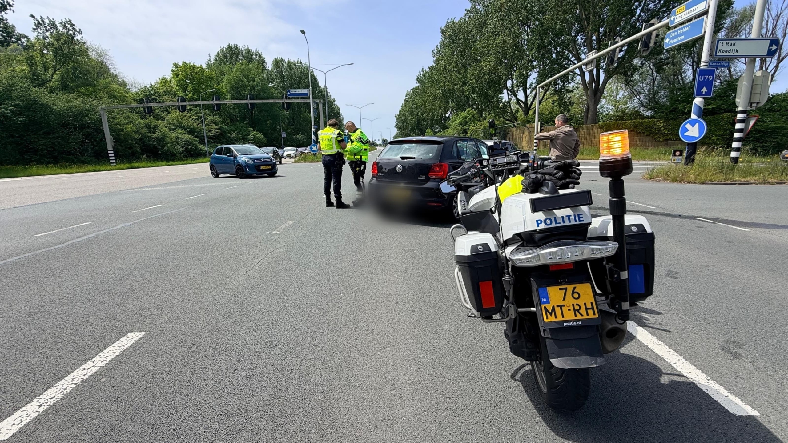 Een zwart voertuig met schade aan de voorkant, omringd door politieauto’s en -motorfiets, met agenten in gesprek op de achtergrond.