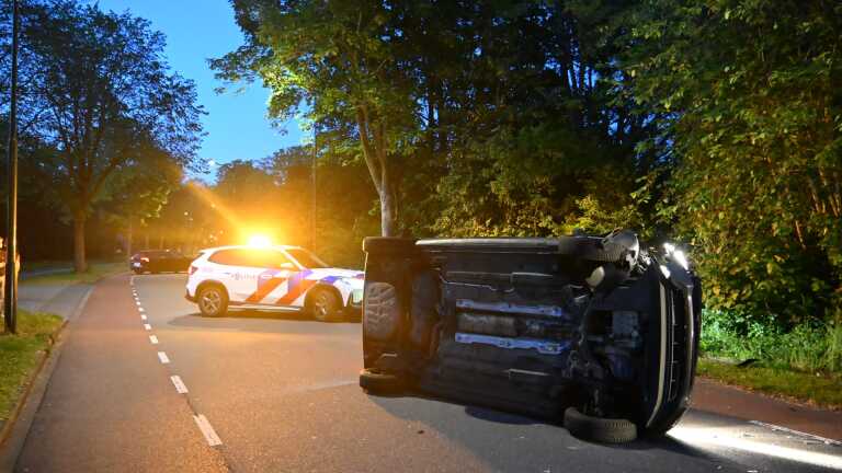 Een gekantelde auto ligt op de weg naast een politieauto met zwaailichten, omgeven door bomen bij schemering.