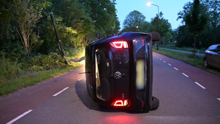 Een gekantelde auto ligt op de weg naast een politieauto met zwaailichten, omgeven door bomen bij schemering.