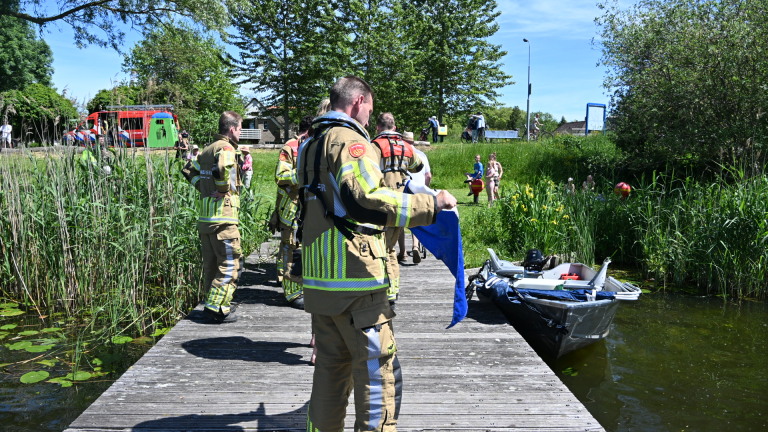 Politie- en brandweermensen staan op een steiger aan een meer, met boten en mensen in de achtergrond.