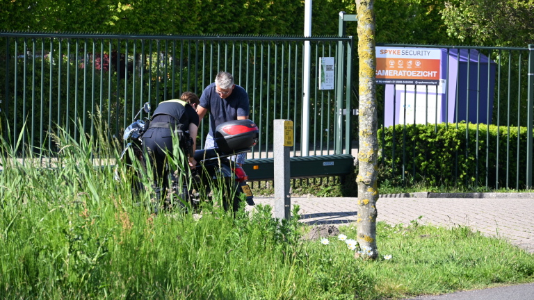 Ambulance en politie bij een incident langs de weg, met een politieagent en een scooter op de voorgrond.