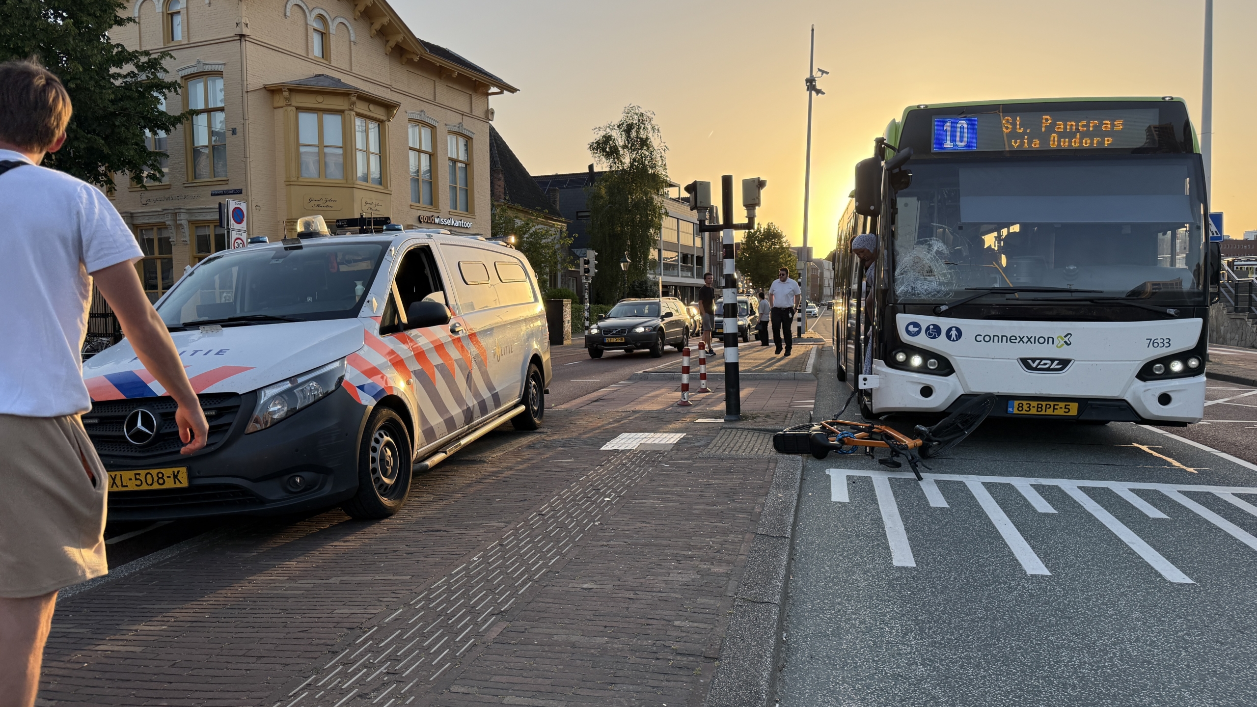 Een bus met een ingedeukte voorruit staat stil op straat met een oranje fiets eronder; een voorbijganger loopt langs.