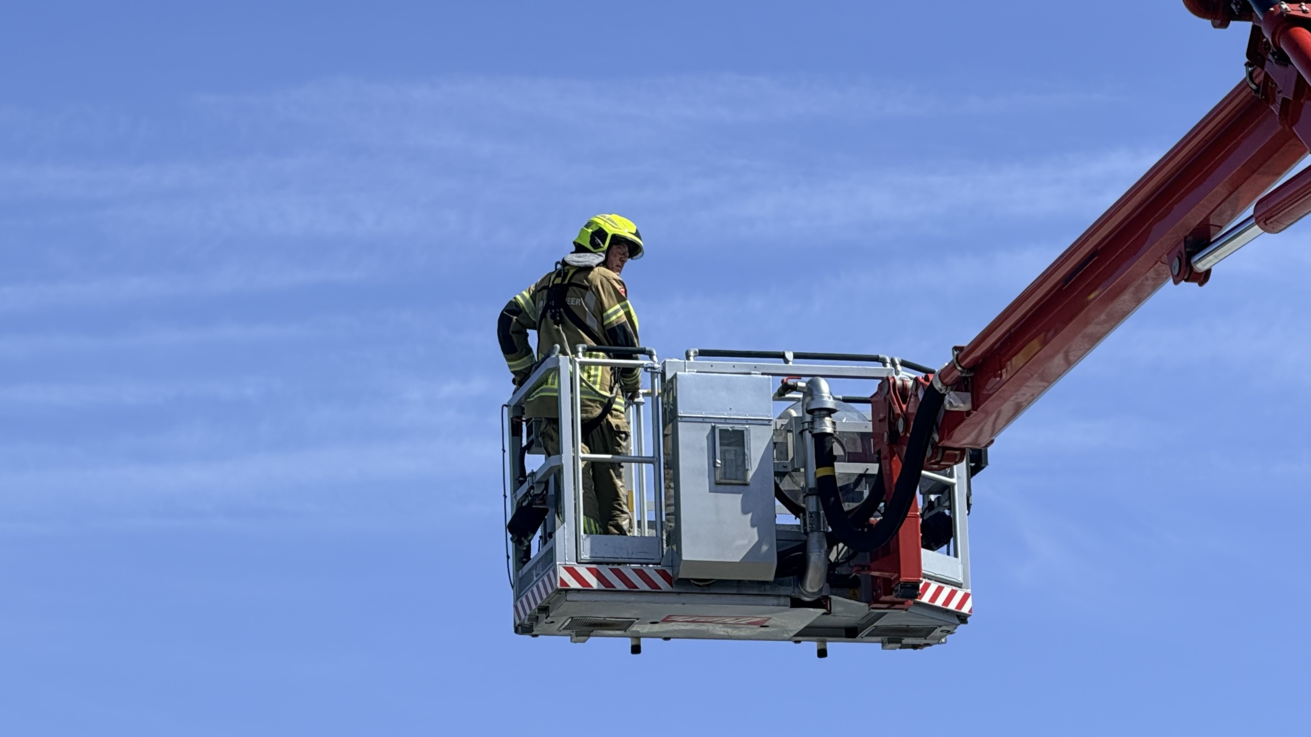 Brandweerlieden in gele helmen werken op een dak met een rode brandslang; een rode brandweerauto staat ernaast.