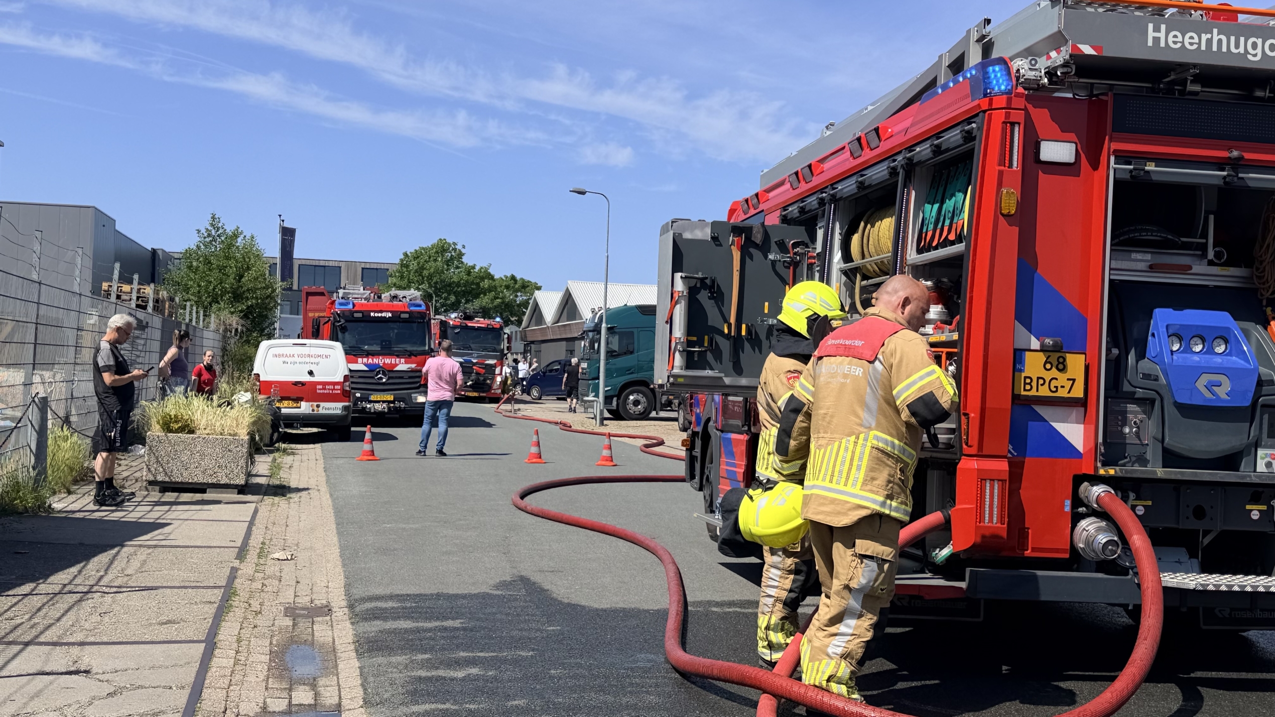 Brandweerlieden in gele helmen werken op een dak met een rode brandslang; een rode brandweerauto staat ernaast.
