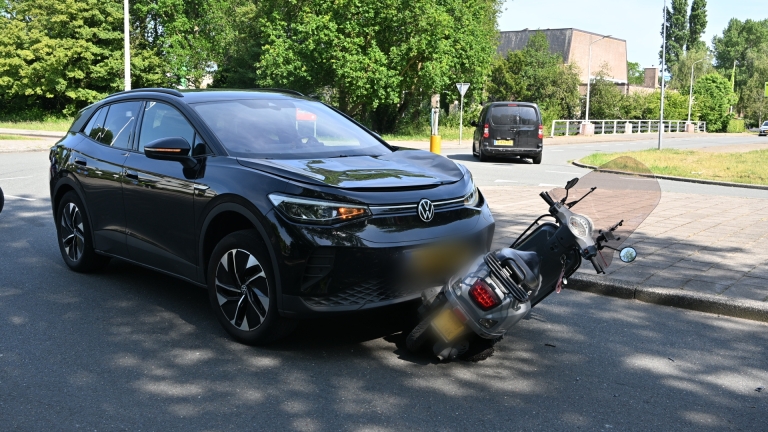Een zwarte auto is botst met een grijze scooter op een straat; een ambulance staat op de achtergrond.