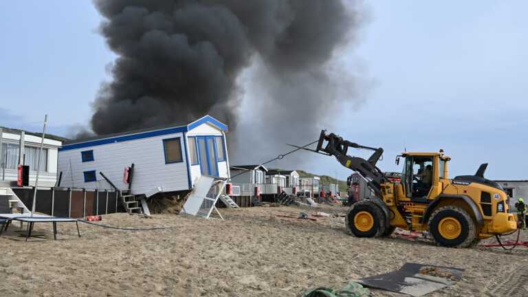 Brandweerlieden bestrijden een uitslaande brand in een strandhuisje, met dikke zwarte rookpluimen stijgend in de lucht.