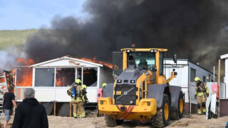 Brandweerlieden bestrijden een uitslaande brand in een strandhuisje, met dikke zwarte rookpluimen stijgend in de lucht.