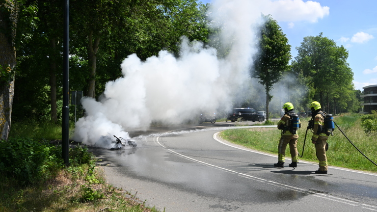 Brandende motorfiets op een weg met dikke zwarte rook, omgeven door groen; enkele toeschouwers op de achtergrond.