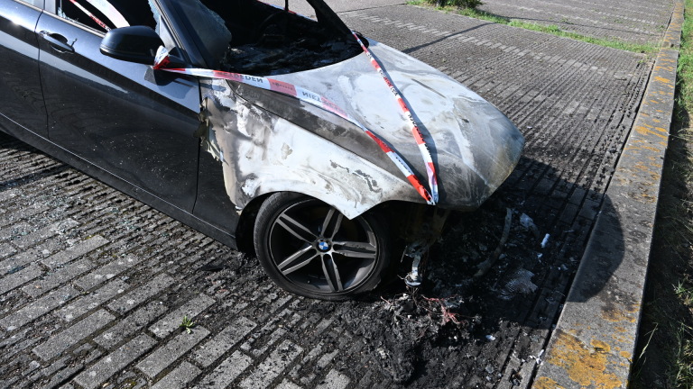 Zwarte auto met brandschade aan de voorzijde, omringd door rood-wit politietape naast een grasveld.