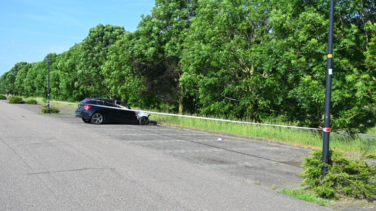 Zwarte auto met brandschade aan de voorzijde, omringd door rood-wit politietape naast een grasveld.