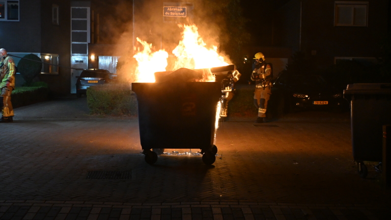Brandende afvalcontainer naast een straat, met vlammen zichtbaar tegen de nachtelijke achtergrond.