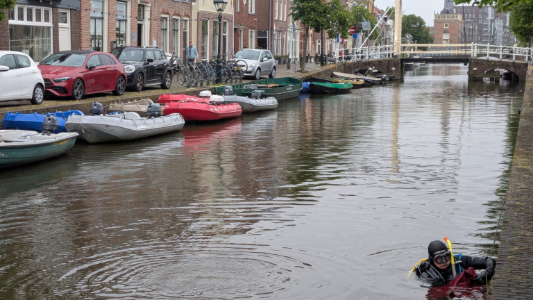 Twee duikers in een kanaal houden een spandoek met een ontwerp en tekst omhoog. Op de achtergrond geparkeerde auto's.