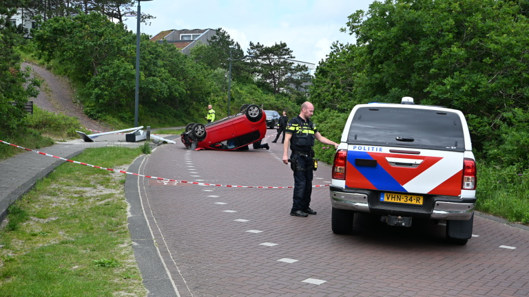 Een rode auto ligt omgekeerd op de weg, met een ambulance en hulpverleners op de achtergrond.