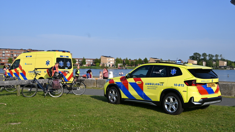 Ambulancemedewerkers staan bij een ambulance op een strand, omringd door mensen en kinderen.