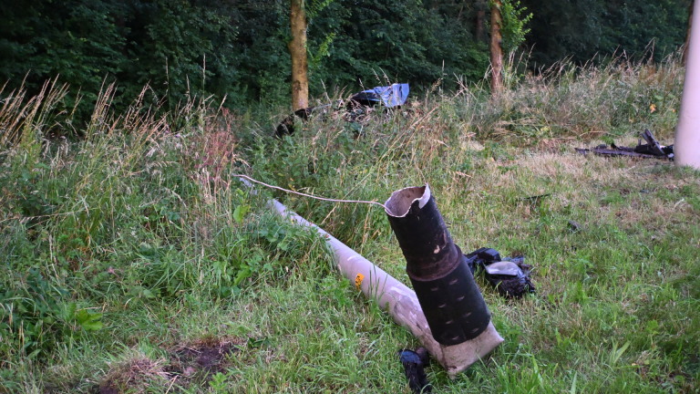 Zwaar beschadigde auto naast de weg in hoog gras, omgeven door bomen.