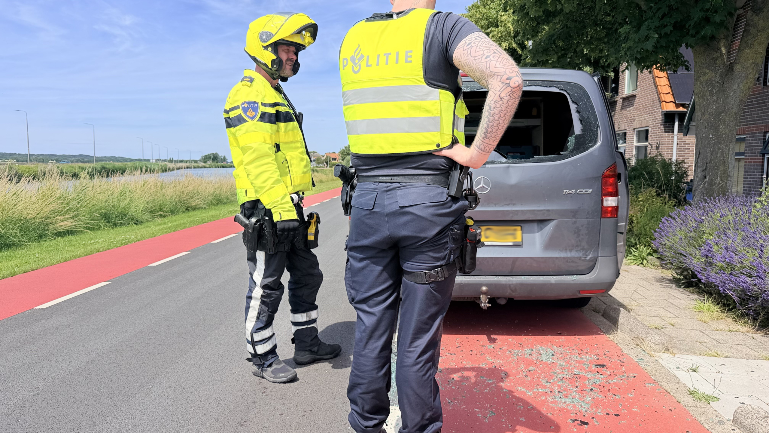 Politieagenten onderzoeken glasscherven op een fietspad naast een geopende bestelwagen, in een landelijke omgeving.