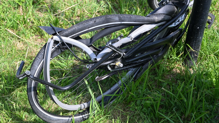 Twee mensen in sportkleding verlenen hulp aan een persoon naast een fiets, met een kijker en een verkeersbord op de achtergrond.