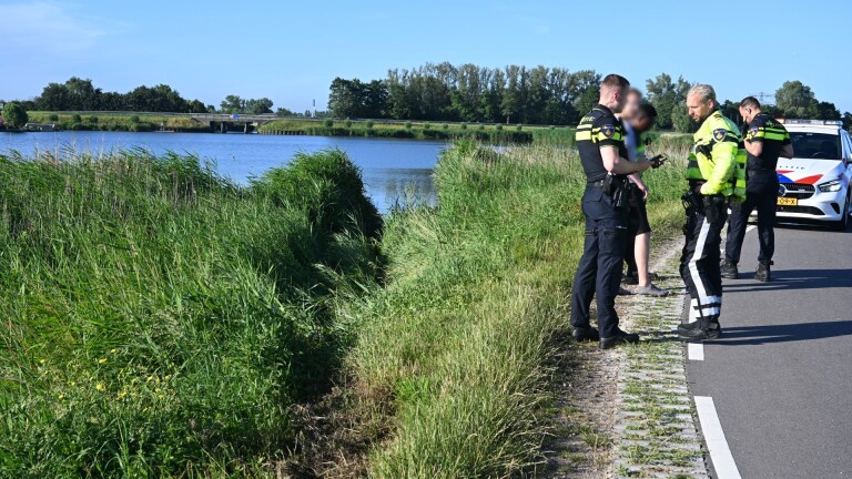 Half gezonken auto in een meer, omgeven door riet en in de verte huizen en een brug.