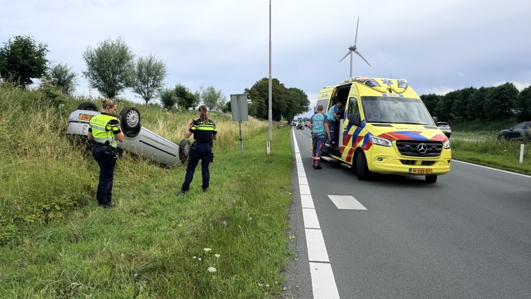 Auto ligt ondersteboven in de berm naast een weg, met politieagenten en een ambulance aanwezig.