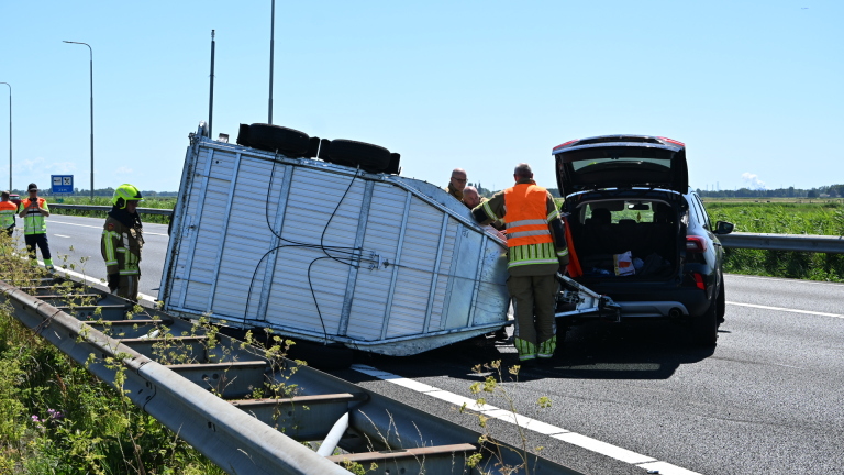 Hulpverleners op de snelweg bij een ongeval, naast een beschadigde auto en een afgebroken aanhanger.