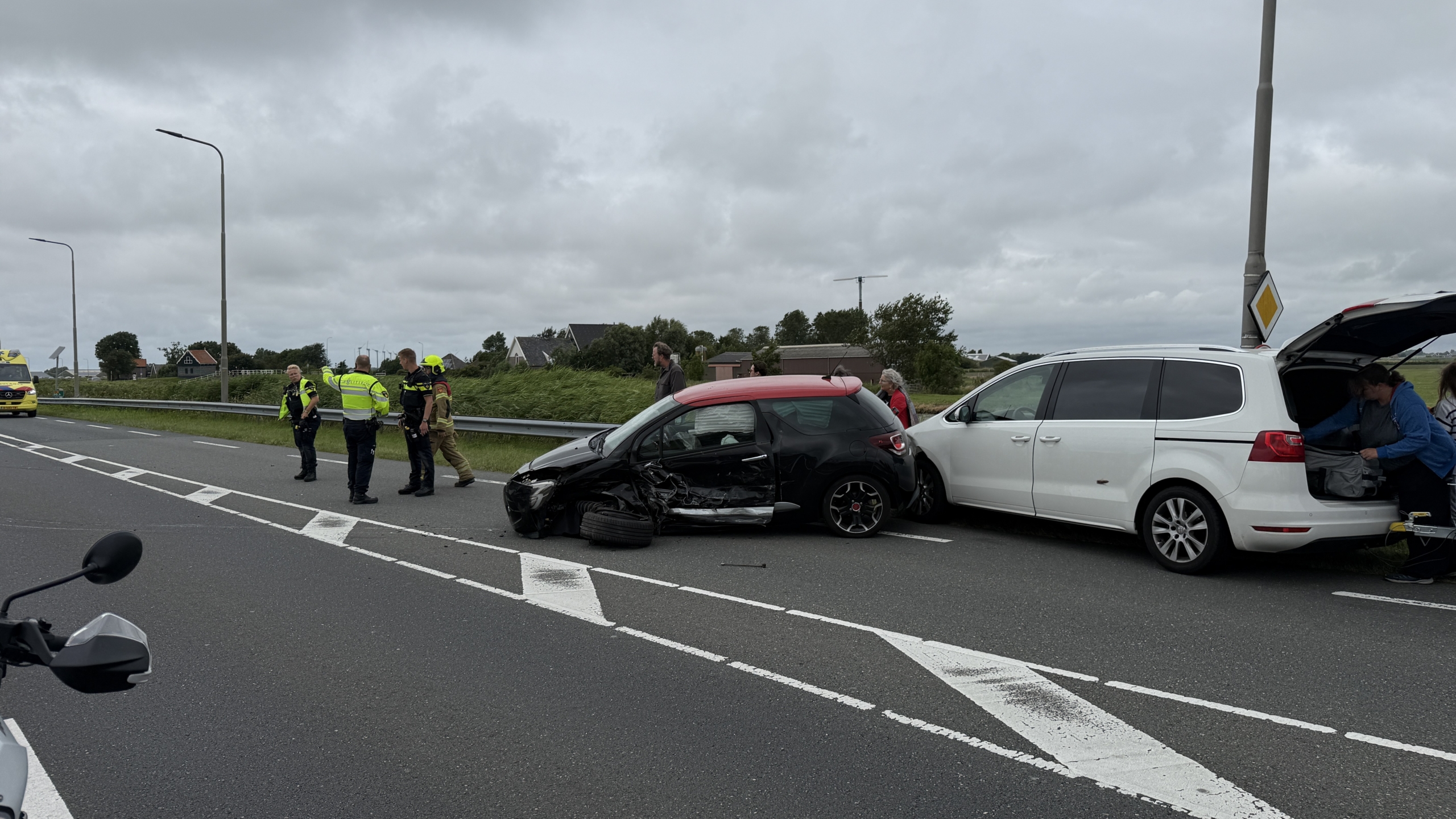 Verkeersongeval op een snelweg met betrokken auto's, brandweerlieden en twee ambulances aanwezig, omstanders kijken toe.