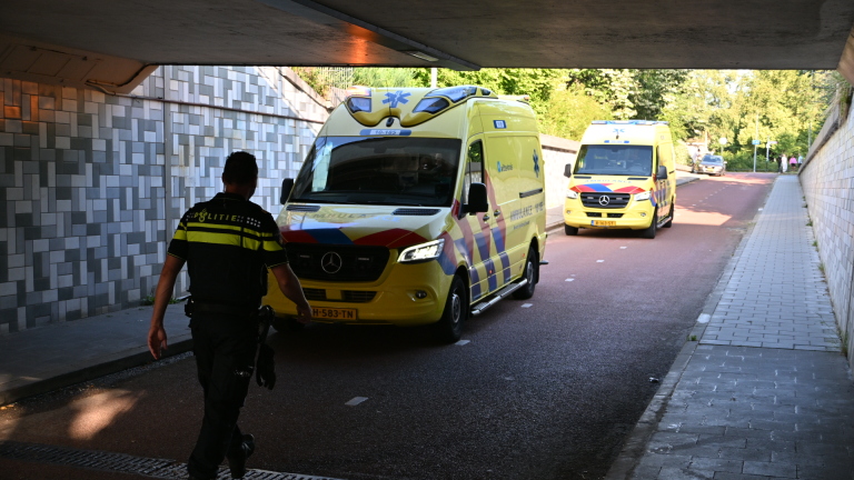 Politieagent loopt in een tunnel langs twee gele ambulances op een fietspad, met daglicht en bomen op de achtergrond