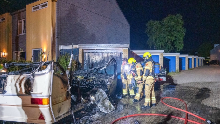 Brandende camper naast een gebouw in de nacht, met hoge vlammen en dikke rookpluim langs een rij garageboxen