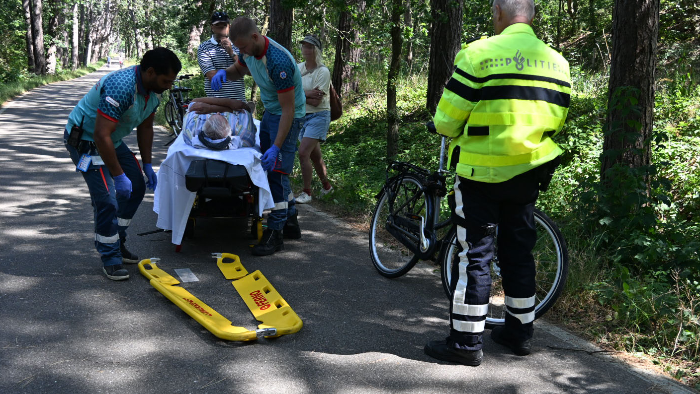 Gele ambulance op een bospad, met fietsers en omstanders eromheen tussen hoge bomen