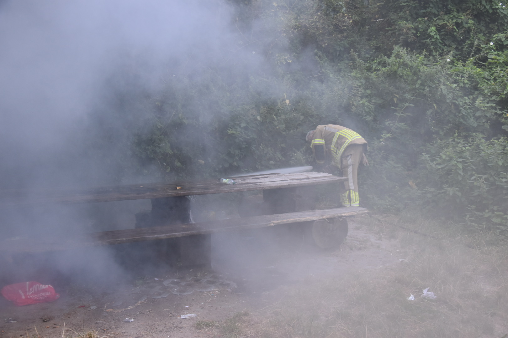Brandweerlieden blussen een smeulende picknicktafel in het bos terwijl rook tussen de bomen hangt