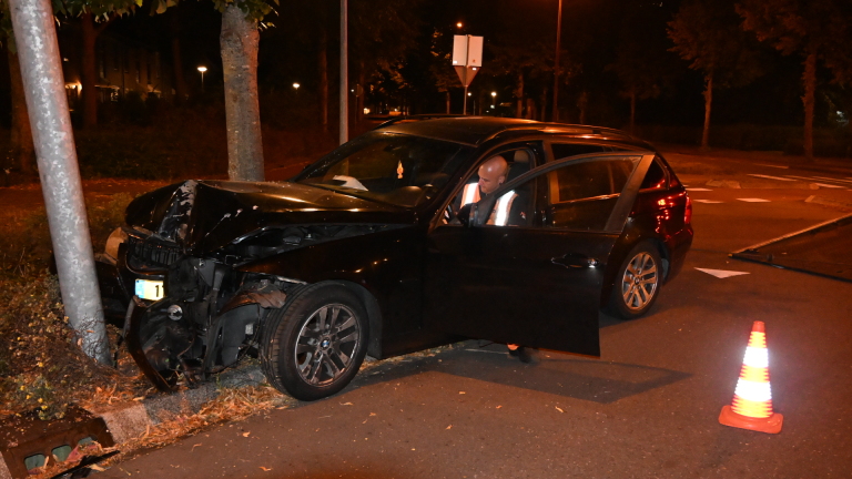 Zwarte auto met zware frontschade tegen een paal, ’s nachts; bestuurder stapt uit, met verkeerskegel op de weg