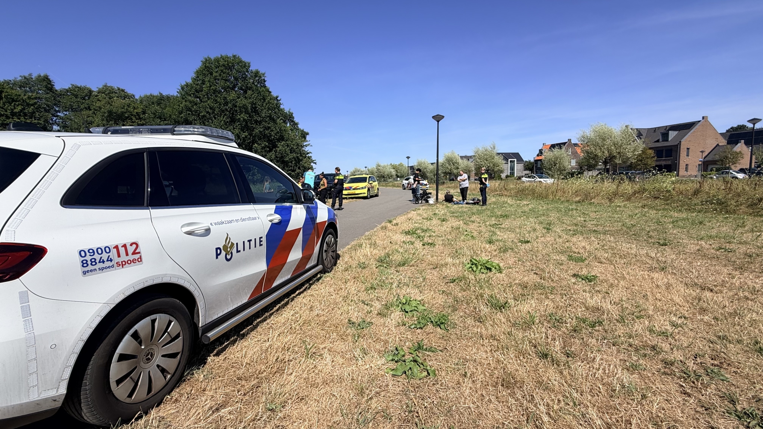 Ambulance met open achterdeuren; hulpverleners behandelen iemand op de grond naast een brancard langs een fietspad
