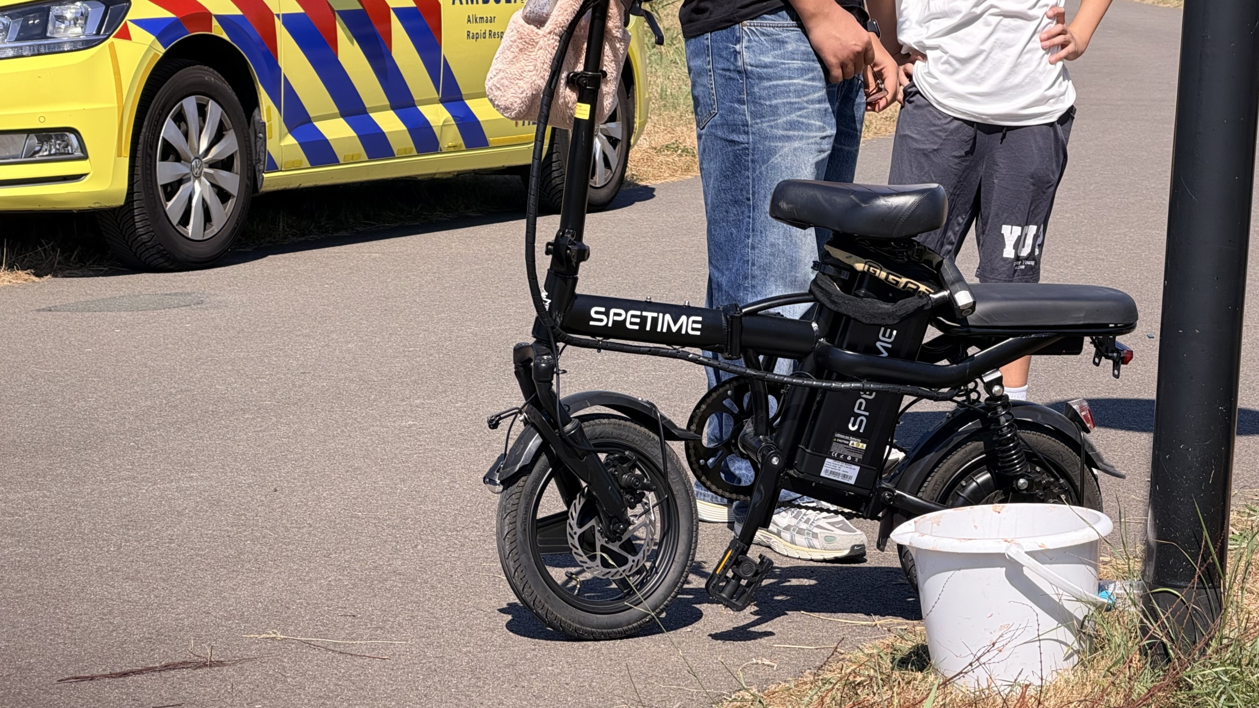 Ambulance met open achterdeuren; hulpverleners behandelen iemand op de grond naast een brancard langs een fietspad