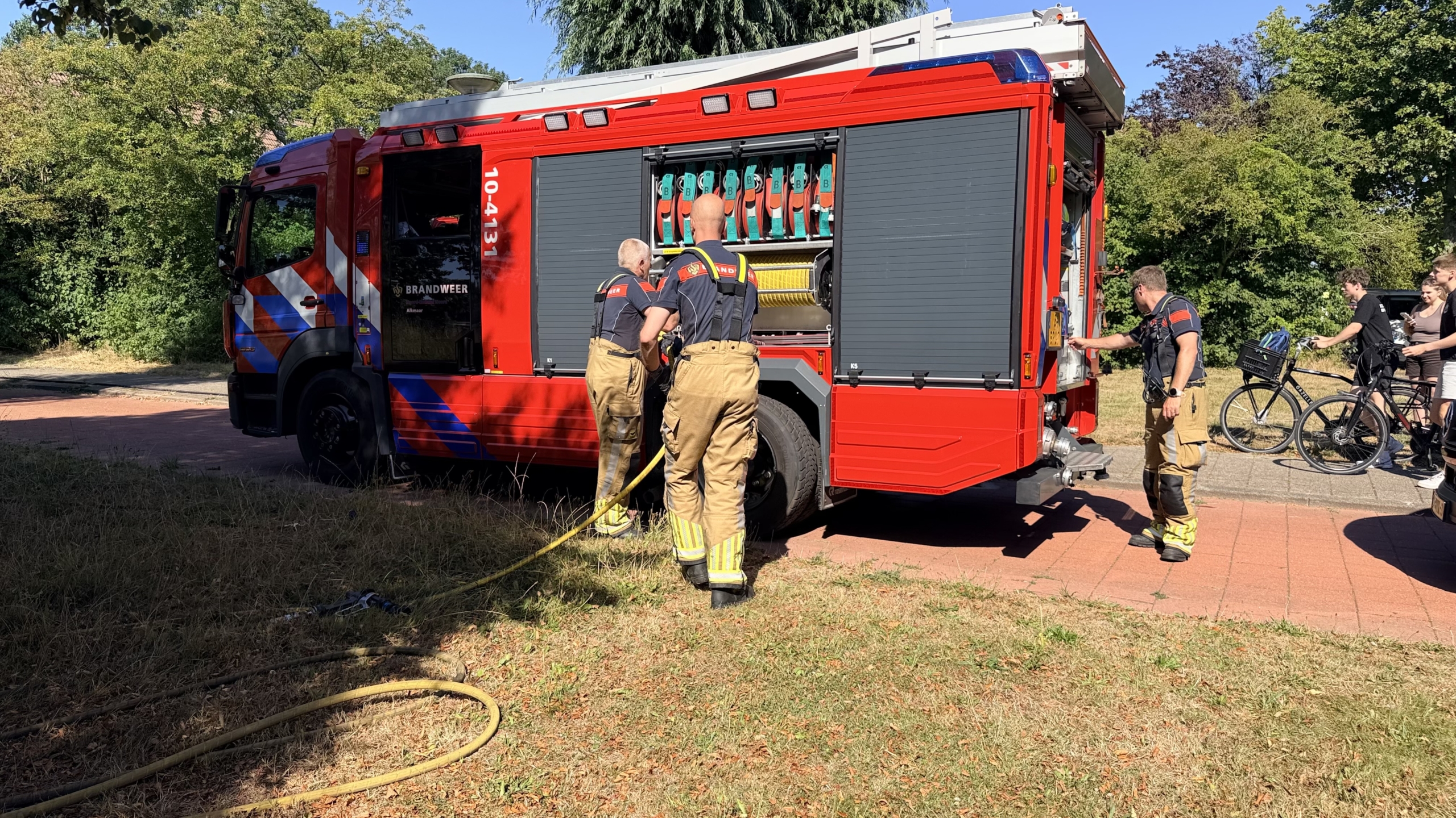 Twee brandweerlieden lopen met slangen over een droog grasveld met rook van een bermbrand, met bomen op de achtergrond