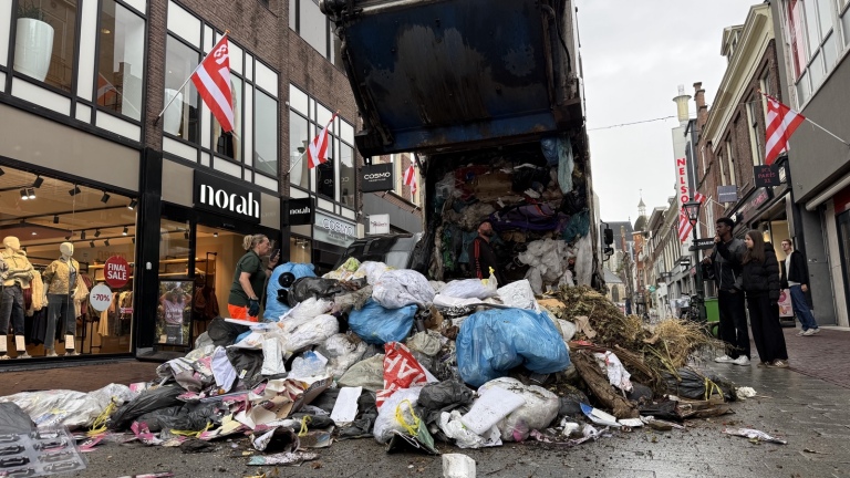 Vuilniswagen lost rokende afvalberg voor de HEMA, terwijl twee medewerkers blussen en omstanders bij de ingang staan