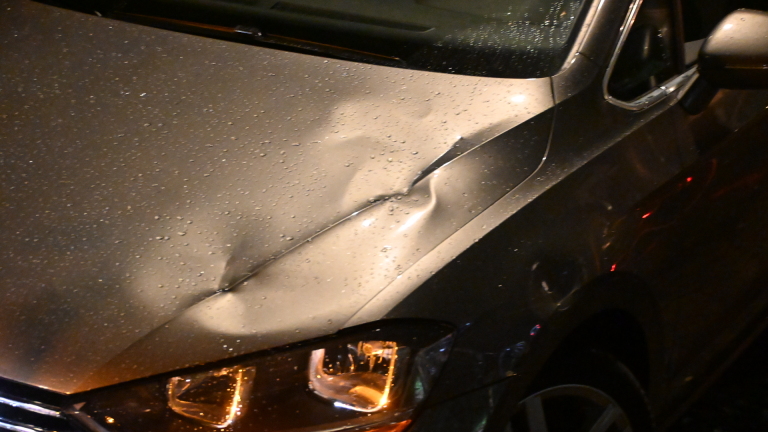 Politieagent telefoneert naast een politieauto op een donkere woonstraat, met een beschadigde paal op de voorgrond