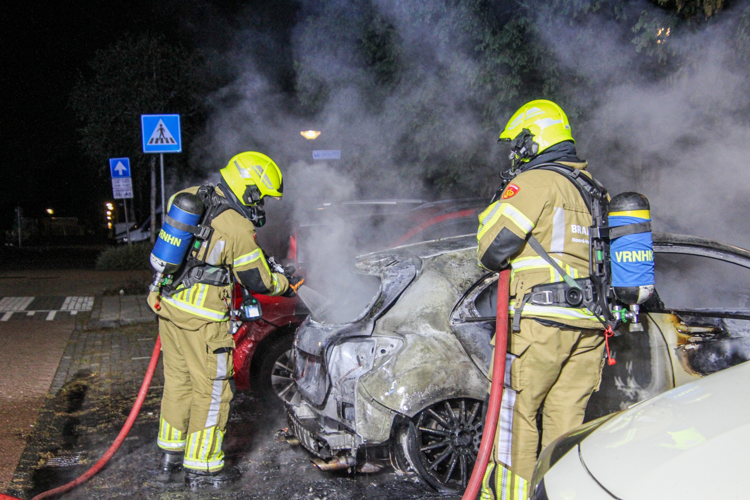Brandweerlieden blussen brandende auto op parkeerplaats, met veel rook tussen geparkeerde auto’s in het donker