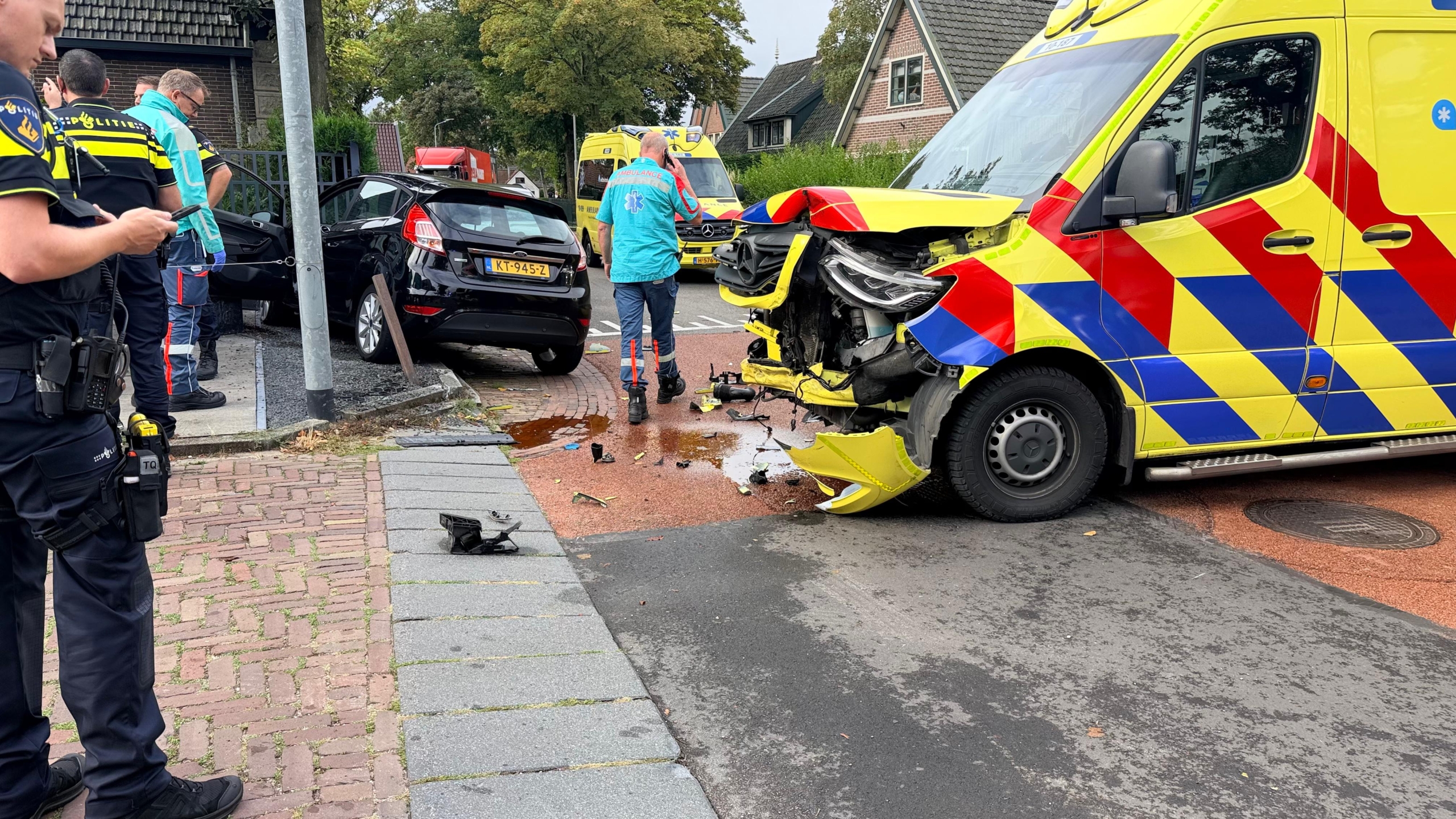 Beschadigde ambulance en politie bij een ongeluk op een kruispunt, met een zwarte auto en brokstukken op de weg