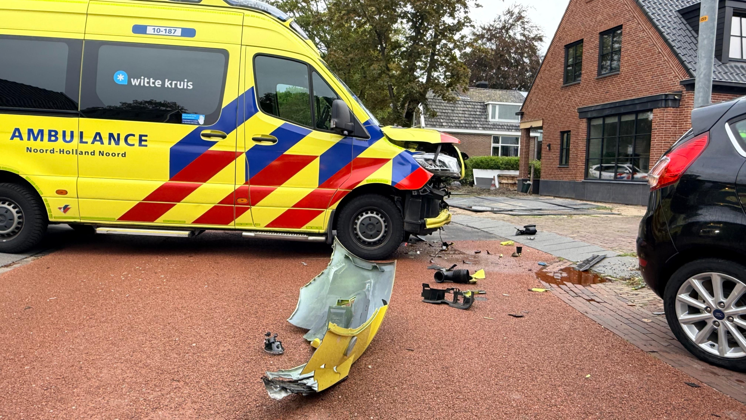 Beschadigde ambulance en politie bij een ongeluk op een kruispunt, met een zwarte auto en brokstukken op de weg