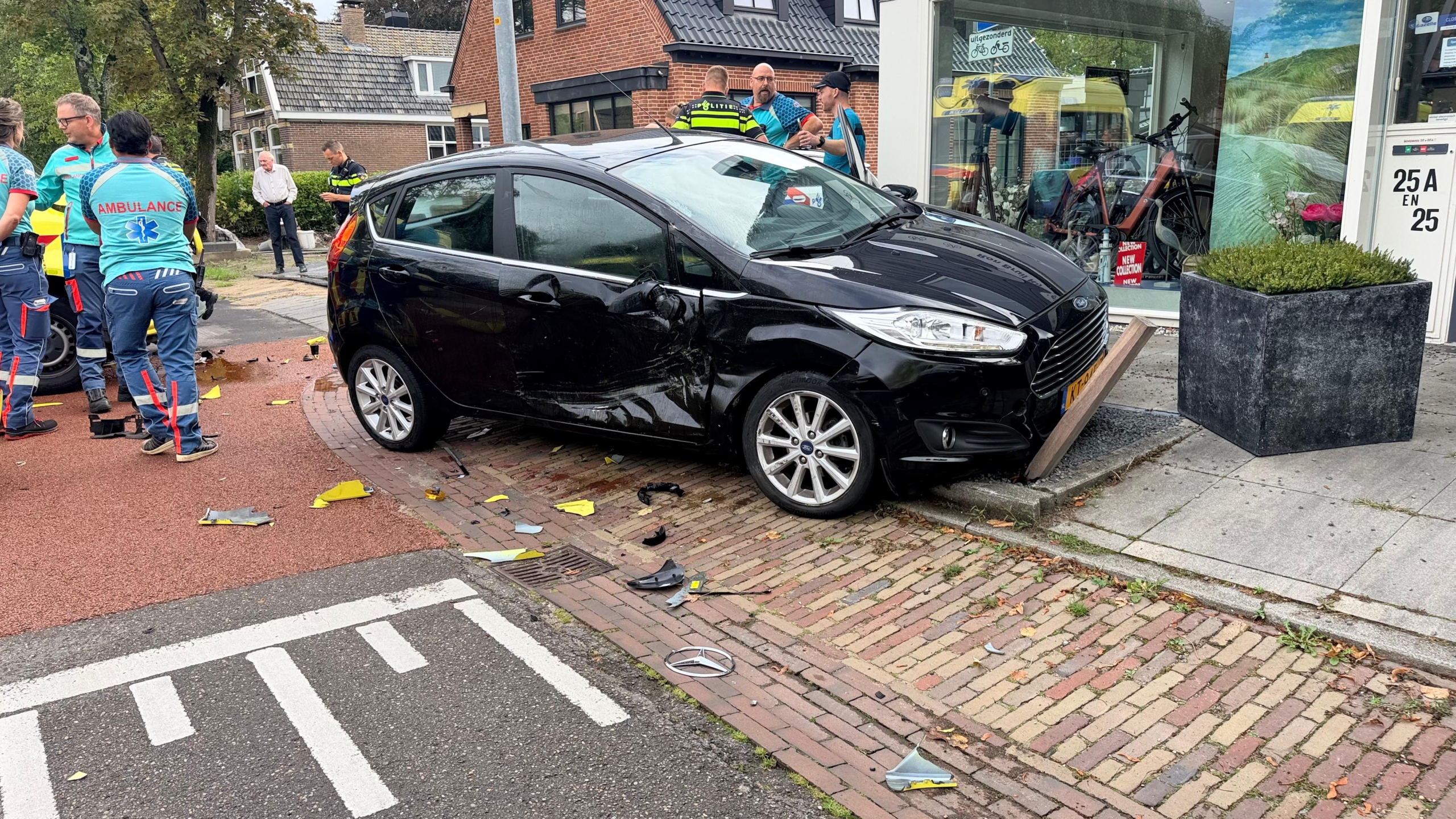 Beschadigde ambulance en politie bij een ongeluk op een kruispunt, met een zwarte auto en brokstukken op de weg