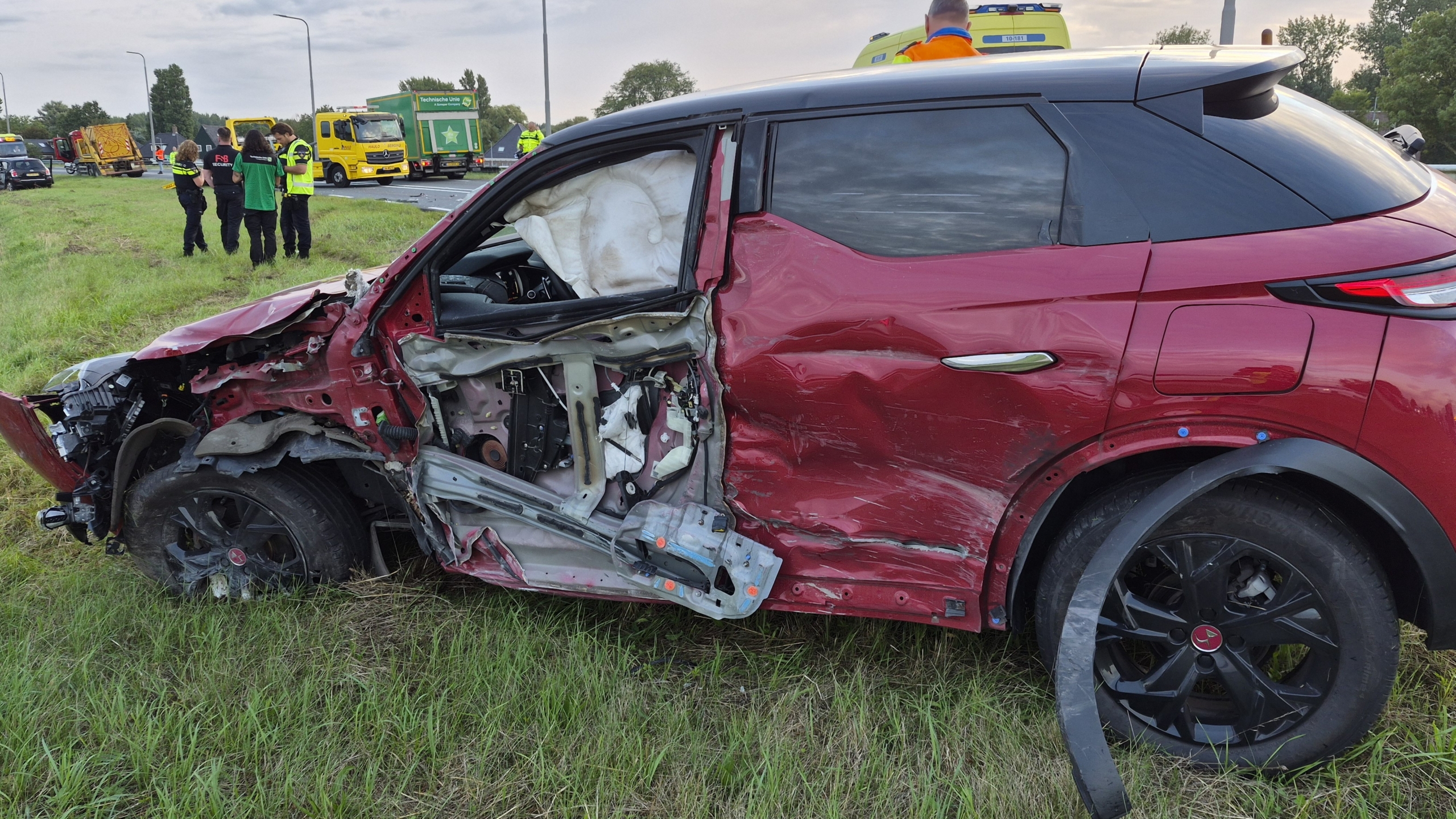 Twee beschadigde auto’s in de berm na aanrijding langs de snelweg, met hulpdiensten en verkeer op de achtergrond