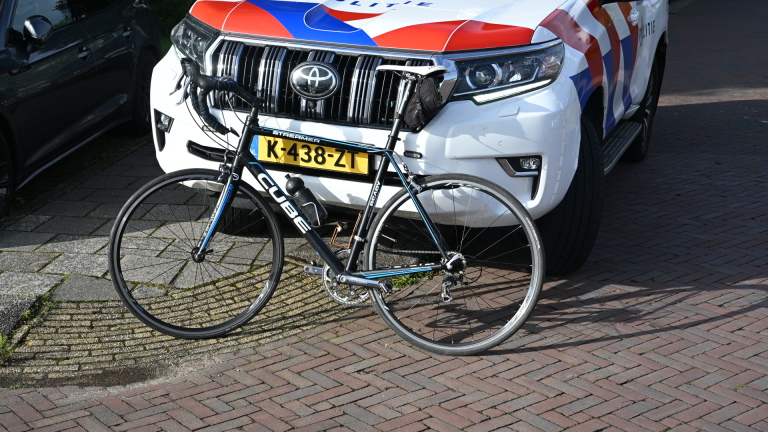 Politieagent en ambulancemedewerker bij blauwe auto met racefiets achterop in parkeervak, met omstanders ernaast