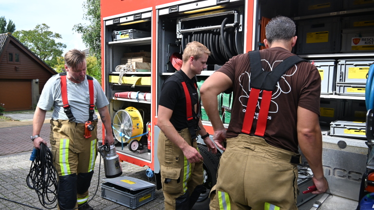 Drie brandweerlieden halen materiaal uit een open brandweerwagen, met slangen, kisten en een ventilator zichtbaar