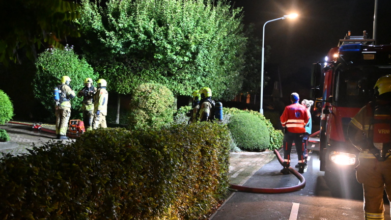 Brandweerlieden met gele helmen overleggen bij een bakstenen gevel, met ademluchtflessen en een officier van dienst
