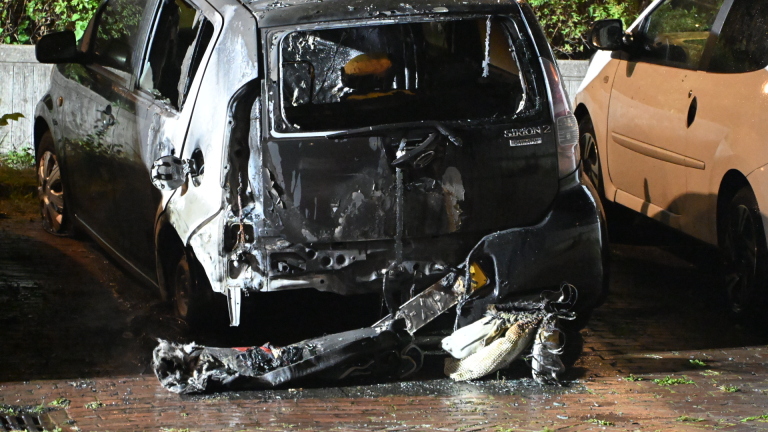 Brandweer blust een brandende auto tussen geparkeerde voertuigen in het donker, met grote vlammen en rook