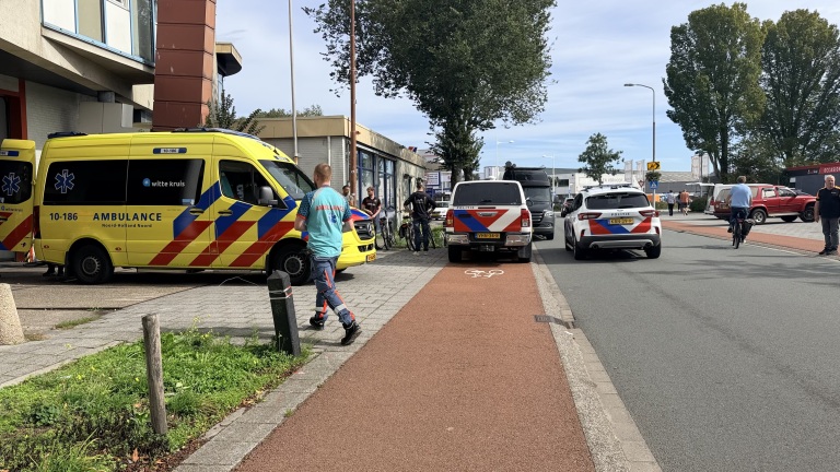 Ambulance en politieauto’s staan langs de weg bij een gebouw, met hulpverlener en omstanders op de stoep