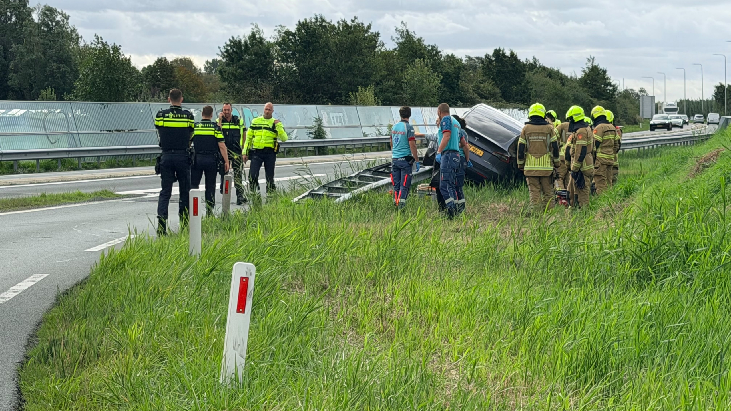Grijze auto staat scheef op de vangrail langs een afrit, met brandweer en ambulance op de achtergrond