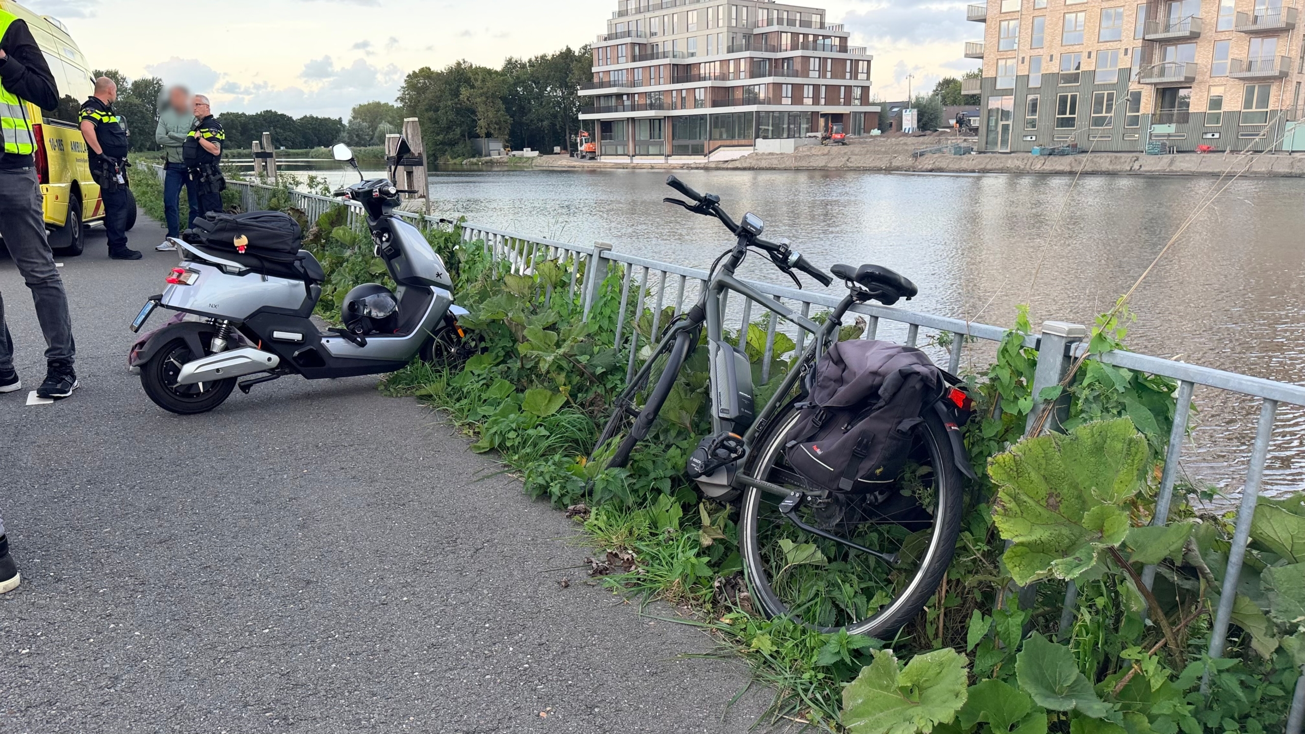 Politieagenten praten naast een geparkeerde ambulance op een fietspad langs het water, met kleding op de grond