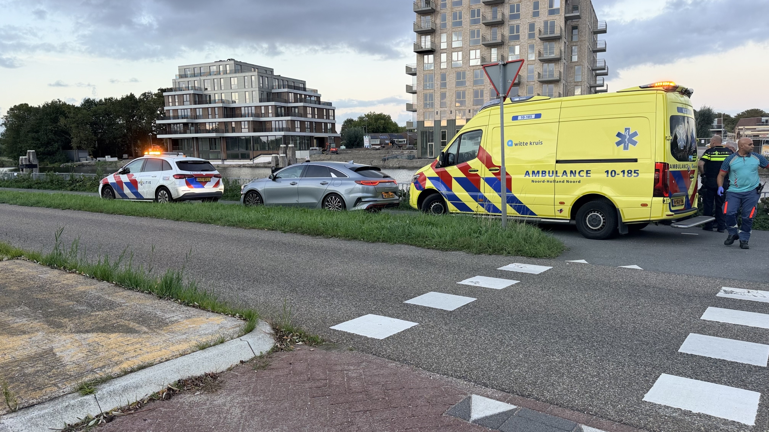 Politieagenten praten naast een geparkeerde ambulance op een fietspad langs het water, met kleding op de grond