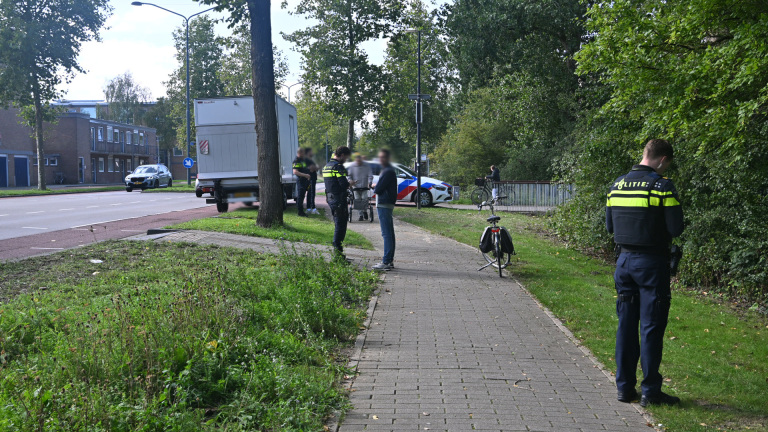 Politieagenten spreken met omstanders naast een geparkeerde vrachtwagen en politieauto op een fietspad langs de weg